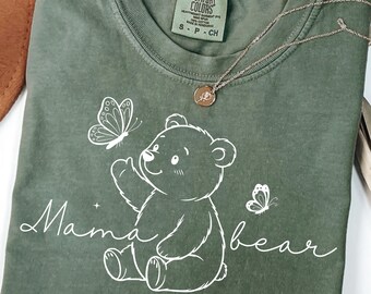 May include: Moss green t-shirt with a white graphic of a bear and butterflies. The text "Mama bear" is written in a script font. A gold necklace is draped across the shirt. The shirt is a Comfort Colors brand.