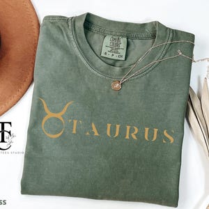 May include: A green t-shirt with a gold Taurus zodiac sign and the word "Taurus" printed on the front.