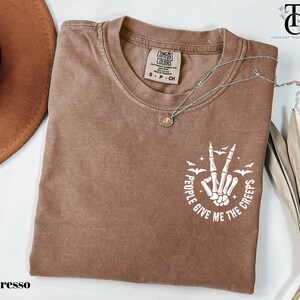 May include: Espresso-colored t-shirt with a crew neck. The shirt features a white graphic of a skeleton hand making a peace sign, surrounded by bats and the text "People Give Me The Creeps". A silver necklace with a pendant is on the shirt.