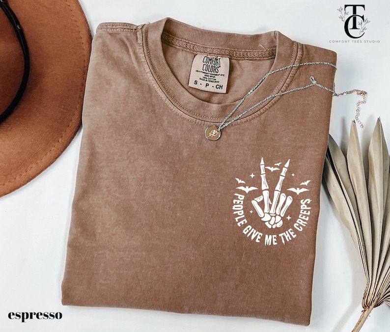 May include: Espresso-colored t-shirt with a crew neck. The shirt features a white graphic of a skeleton hand making a peace sign, surrounded by bats and the text "People Give Me The Creeps". A silver necklace with a pendant is on the shirt.