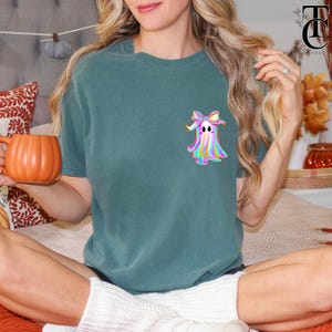 May include: Teal-colored t-shirt featuring a colorful ghost design with a bow. The ghost has a rainbow-colored body and a cute expression. The shirt is a comfortable, casual style, perfect for Halloween or everyday wear.