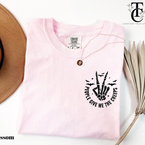 May include: A light pink t-shirt with the text "I GIVE ME THE CREEPS" in a circular design with a skeleton hand making a peace sign. The shirt also features small bat graphics. The shirt is a Comfort Colors brand.