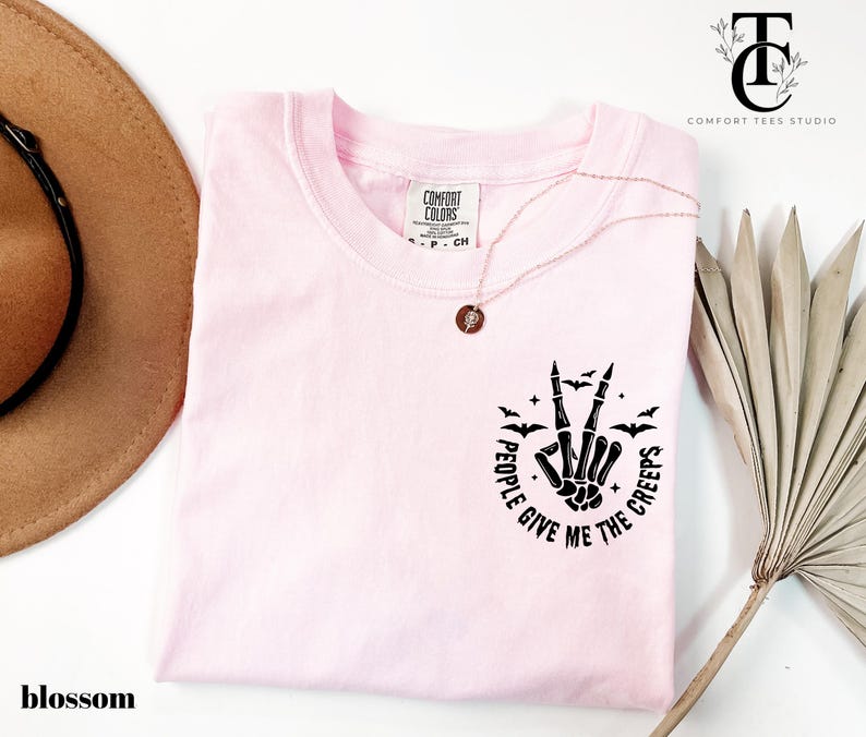 May include: A light pink t-shirt with the text "I GIVE ME THE CREEPS" in a circular design with a skeleton hand making a peace sign. The shirt also features small bat graphics. The shirt is a Comfort Colors brand.