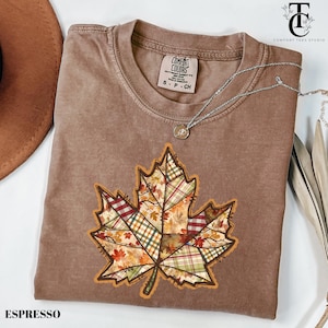 May include: Espresso-coloured t-shirt with a large, patchwork leaf design. The leaf features various patterns and colours, including plaid and floral prints. A silver necklace with a pendant rests on the shirt. The shirt has a Comfort Colors tag.
