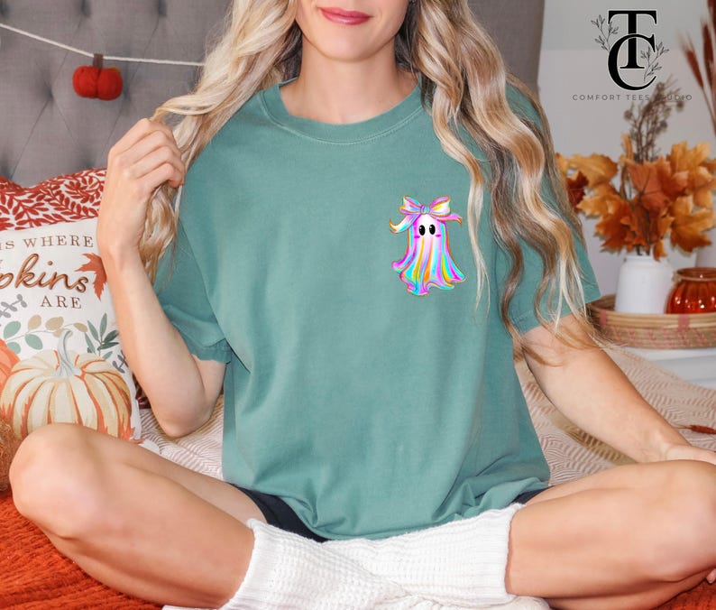 May include: A teal-colored t-shirt featuring a colorful ghost graphic with a bow. The ghost design has a rainbow color scheme. The shirt is a crew neck style and appears to be made of a soft material. The shirt is part of a Halloween or fall-themed collection.