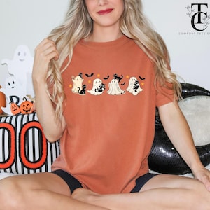 Op de afbeelding: Roestkleurig T-shirt met een Halloween-thema. Het ontwerp bevat vier cartoongeesten, elk met een zwarte kat, vleermuizen en sterren. Het shirt maakt deel uit van de Comfort Tees Studio collectie.