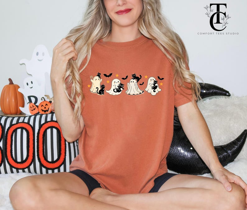 Op de afbeelding: Roestkleurig T-shirt met een Halloween-thema. Het ontwerp bevat vier cartoongeesten, elk met een zwarte kat, vleermuizen en sterren. Het shirt maakt deel uit van de Comfort Tees Studio collectie.
