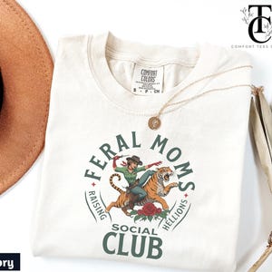 May include: Ivory t-shirt with a green circle graphic that says "Feral Moms Raising Hellions Social Club". Inside the circle is a cartoon image of a woman riding a tiger.