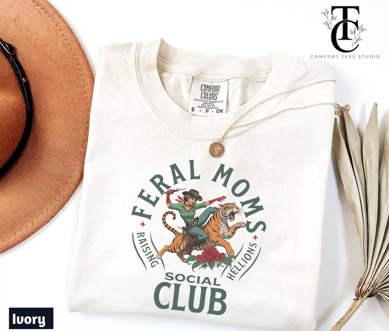 May include: Ivory t-shirt with a green circle graphic that says "Feral Moms Raising Hellions Social Club". Inside the circle is a cartoon image of a woman riding a tiger.