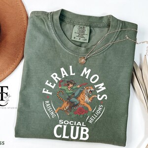 May include: A green t-shirt with a white graphic design that says "Feral Moms Raising Hellions Social Club". The graphic features a woman riding a tiger.