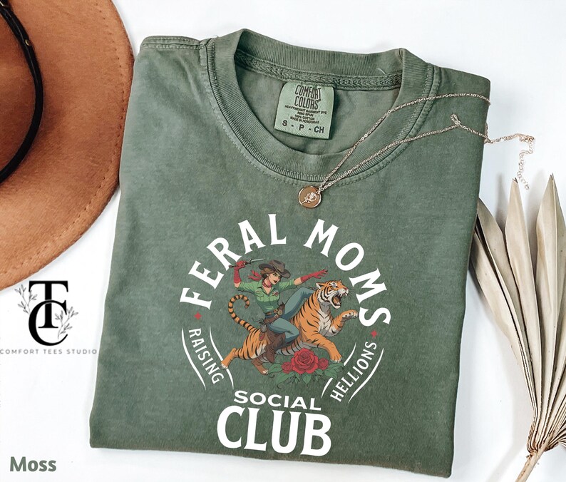 May include: A green t-shirt with a white graphic design that says "Feral Moms Raising Hellions Social Club". The graphic features a woman riding a tiger.