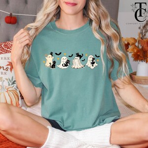 Op de afbeelding: Tealkleurig T-shirt met een Halloween-thema. Het ontwerp bevat vier cartoongeesten, elk vergezeld van een zwarte kat, en verschillende zwarte vleermuizen. Het shirt is een comfortabele, casual stijl, perfect voor het herfstseizoen.