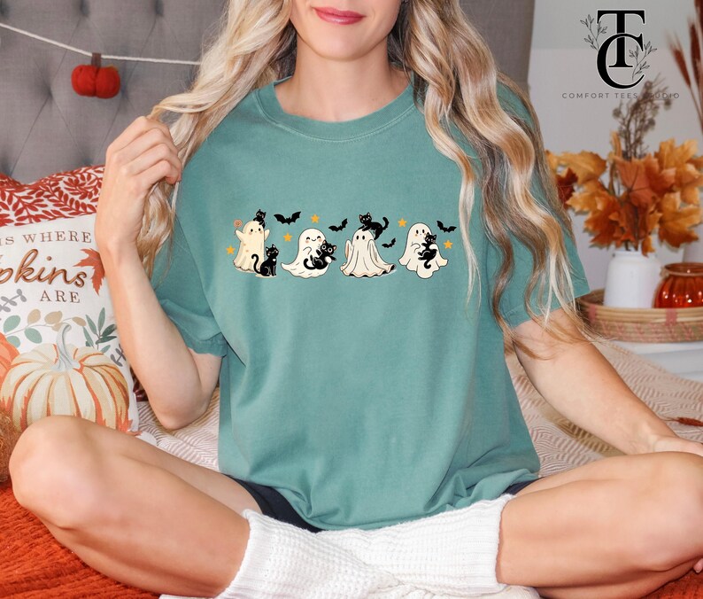 Op de afbeelding: Tealkleurig T-shirt met een Halloween-thema. Het ontwerp bevat vier cartoongeesten, elk vergezeld van een zwarte kat, en verschillende zwarte vleermuizen. Het shirt is een comfortabele, casual stijl, perfect voor het herfstseizoen.