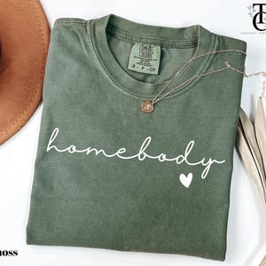 May include: A moss green t-shirt with the text "homebody" in white cursive lettering and a small white heart. The shirt is folded and laid flat on a white surface.