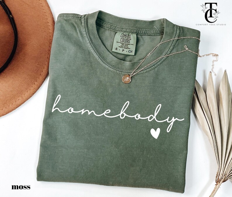 May include: A moss green t-shirt with the text "homebody" in white cursive lettering and a small white heart. The shirt is folded and laid flat on a white surface.