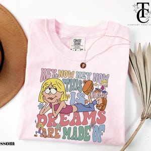 May include: Light pink t-shirt with a graphic featuring a cartoon character and the text "HEY NOW HEY NOW THIS IS DREAMS ARE MADE OF". The shirt has a crew neck and short sleeves. A gold necklace is draped across the shirt.
