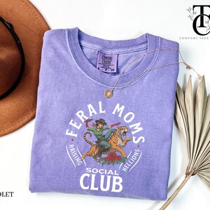 May include: A purple t-shirt with a graphic of a woman riding a tiger. The graphic is surrounded by a circle with the text "Feral Moms Social Club Raising Hellions".