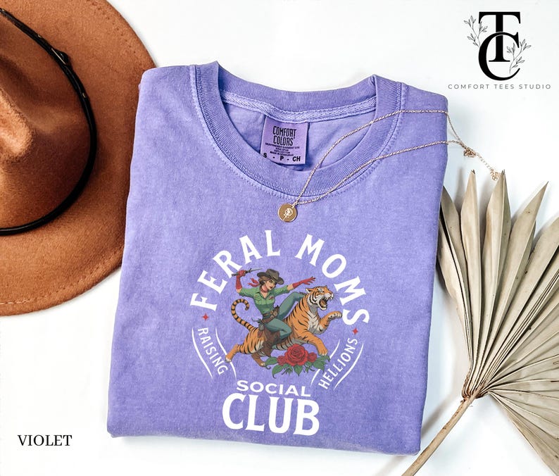 May include: A purple t-shirt with a graphic of a woman riding a tiger. The graphic is surrounded by a circle with the text "Feral Moms Social Club Raising Hellions".