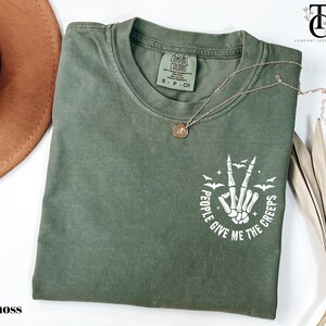 May include: Moss green t-shirt with a crew neck and short sleeves. The shirt features a white graphic of a skeleton hand making a peace sign, surrounded by bats and the text "PEOPLE GIVE ME THE CREEPS". A gold necklace is draped across the shirt.