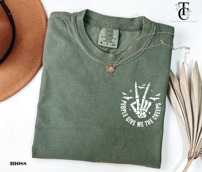 May include: Moss green t-shirt with a crew neck and short sleeves. The shirt features a white graphic of a skeleton hand making a peace sign, surrounded by bats and the text "PEOPLE GIVE ME THE CREEPS". A gold necklace is draped across the shirt.