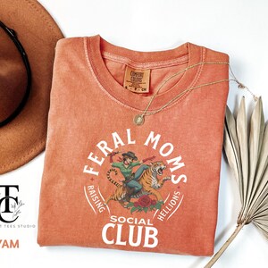 May include: A coral orange t-shirt with a white graphic design that says "Feral Moms Social Club Raising Helllions". The graphic features a woman riding a tiger.