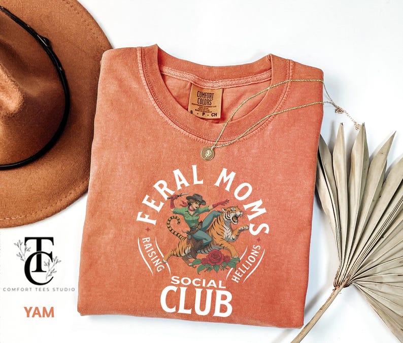 May include: A coral orange t-shirt with a white graphic design that says "Feral Moms Social Club Raising Helllions". The graphic features a woman riding a tiger.