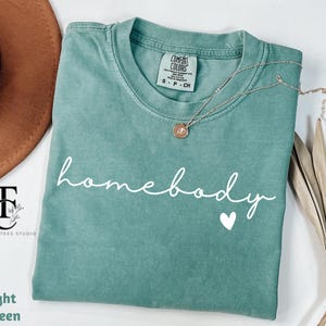 May include: A light green t-shirt with the text "homebody" in white cursive lettering and a small white heart below the text.