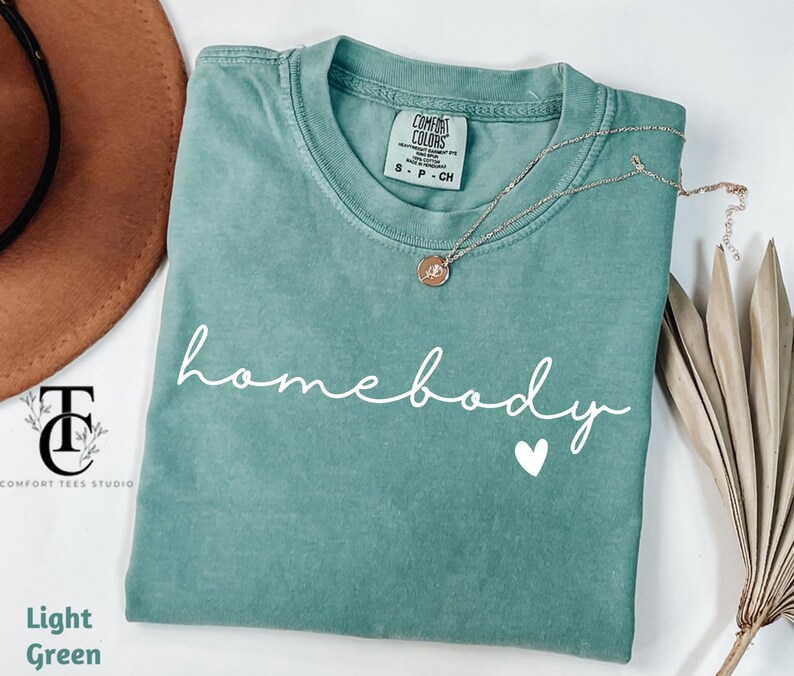 May include: A light green t-shirt with the text "homebody" in white cursive lettering and a small white heart below the text.