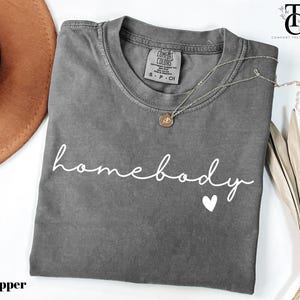 May include: A grey t-shirt with the text "homebody" in white script with a small white heart underneath. The shirt is folded and laid flat on a white surface.