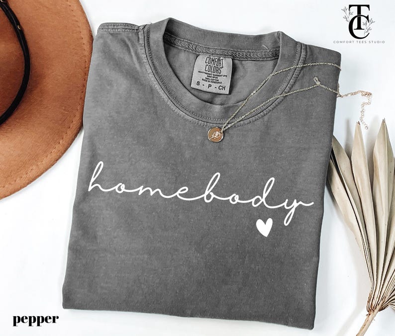 May include: A grey t-shirt with the text "homebody" in white script with a small white heart underneath. The shirt is folded and laid flat on a white surface.