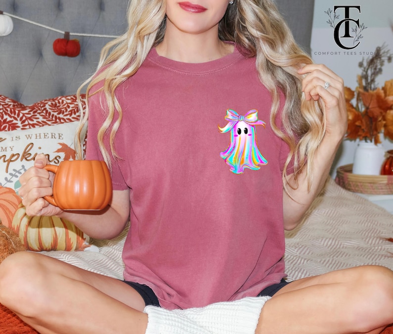 May include: A maroon t-shirt featuring a colorful ghost design with a bow. The ghost has a rainbow-colored body and a cute expression. The person is holding an orange pumpkin-shaped mug, perfect for the fall season.