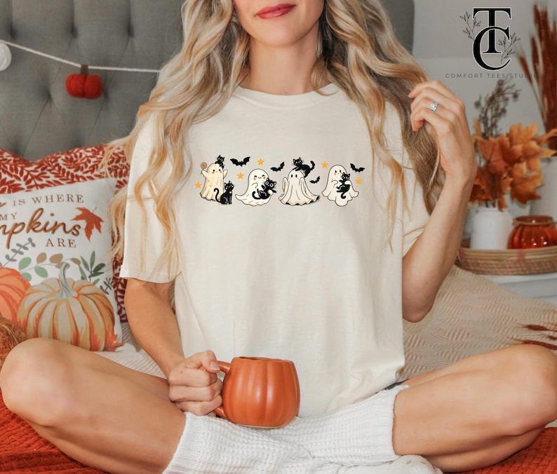 Op de afbeelding: Cr&egrave;mekleurig T-shirt met een Halloween-thema. Het ontwerp bevat cartoongeesten, zwarte katten en sterren. Het shirt wordt gecombineerd met een pompoenvormige mok. De tekst "Comfort Tee Studio" is zichtbaar.