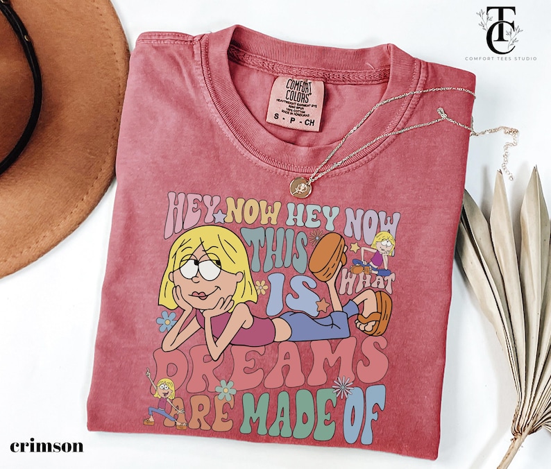 May include: Crimson Comfort Colors t-shirt with a graphic of a cartoon character and the text "HEY NOW HEY NOW THIS IS WHAT DREAMS ARE MADE OF". The shirt has a crew neck and short sleeves. A silver necklace with a pendant is on the shirt.