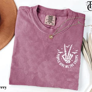 May include: Berry-colored t-shirt with a crew neck and short sleeves. The shirt features a white graphic of a skeleton hand making a peace sign, with the text "PEOPLE GIVE ME THE CREEPS". A gold necklace is draped across the shirt.