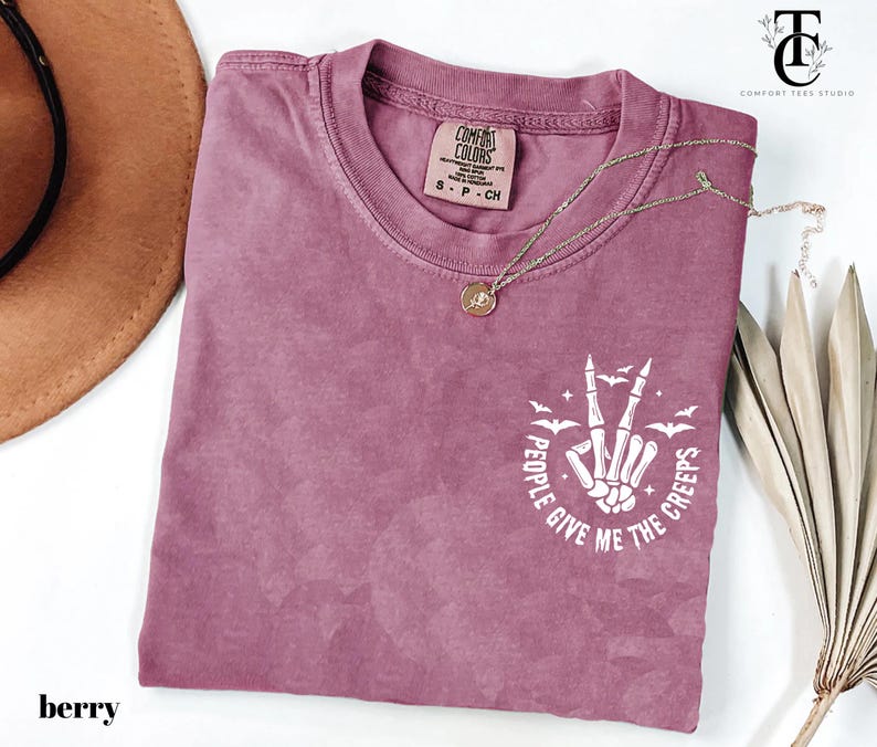 May include: Berry-colored t-shirt with a crew neck and short sleeves. The shirt features a white graphic of a skeleton hand making a peace sign, with the text "PEOPLE GIVE ME THE CREEPS". A gold necklace is draped across the shirt.