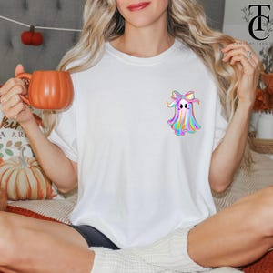 May include: White t-shirt featuring a colorful ghost design with a bow. The ghost has a rainbow color scheme. The person is holding an orange pumpkin-shaped mug. The shirt is part of a fall-themed collection.