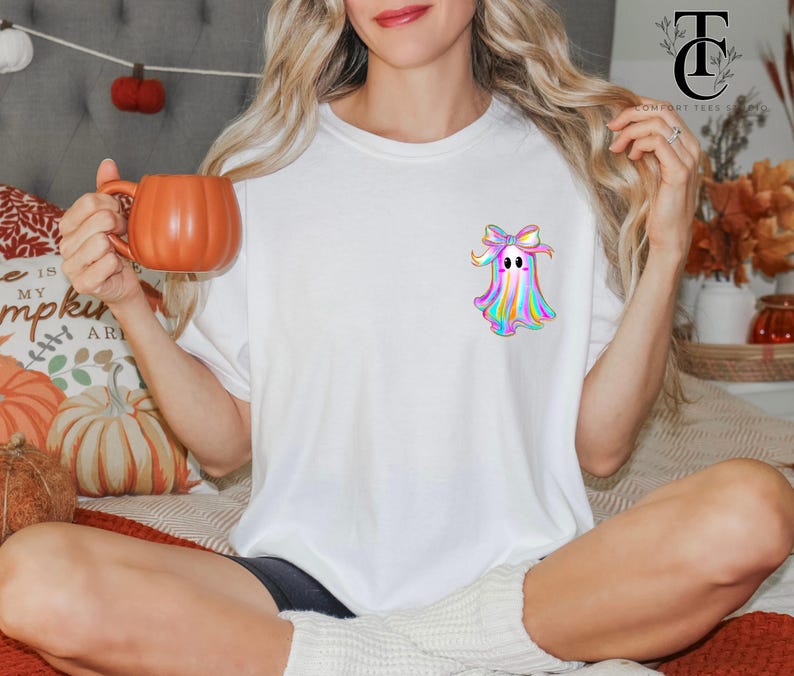 May include: White t-shirt featuring a colorful ghost design with a bow. The ghost has a rainbow color scheme. The person is holding an orange pumpkin-shaped mug. The shirt is part of a fall-themed collection.