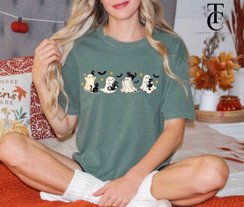 Op de afbeelding: Groen t-shirt met een Halloween-ontwerp. Het ontwerp toont vier cartoongeesten, zwarte katten, vleermuizen en sterren. Het Comfort Studio-logo is zichtbaar. Het shirt is een casual, comfortabele stijl.