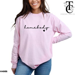 May include: A pink long-sleeved t-shirt with the text "homebody" in black script and a small black heart.