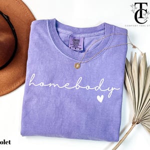 May include: A purple t-shirt with the text "homebody" in white script with a small white heart below. The shirt is made of a soft, comfortable material.
