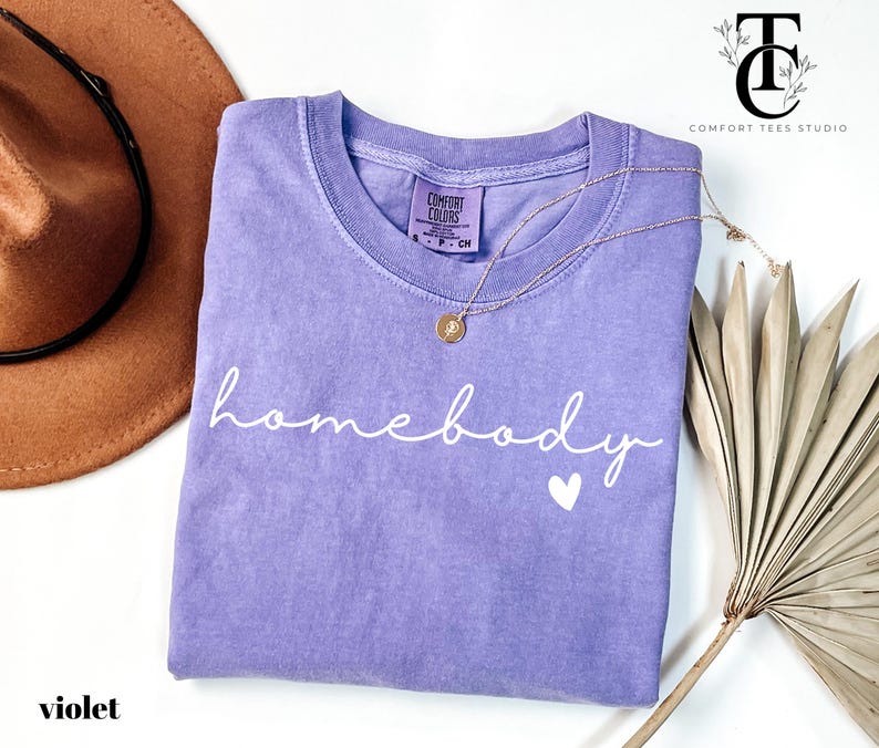 May include: A purple t-shirt with the text "homebody" in white script with a small white heart below. The shirt is made of a soft, comfortable material.