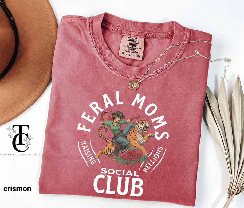 May include: A red t-shirt with a white graphic design that says 'Feral Moms Social Club Raising Hellions'. The graphic also features a cartoon image of a woman riding a tiger. The t-shirt is made of a soft, comfortable fabric and is perfect for everyday wear.