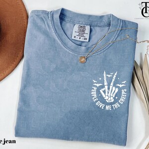 May include: Blue jean colored t-shirt with a crew neck. The shirt features a white graphic of a skeleton hand making a peace sign, with the text "People Give Me The Creeps". A gold necklace is draped across the shirt.