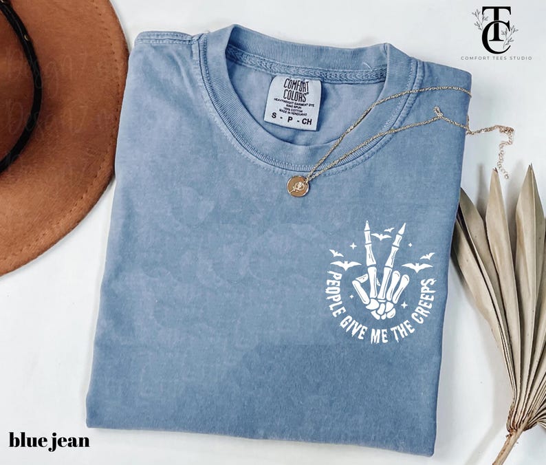 May include: Blue jean colored t-shirt with a crew neck. The shirt features a white graphic of a skeleton hand making a peace sign, with the text "People Give Me The Creeps". A gold necklace is draped across the shirt.