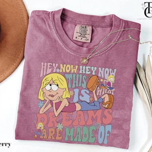 May include: Berry-colored t-shirt with a graphic of a cartoon character and the text "HEY NOW HEY NOW THIS IS WHAT DREAMS ARE MADE OF." The shirt has a crew neck and short sleeves. A gold necklace is draped across the shirt.