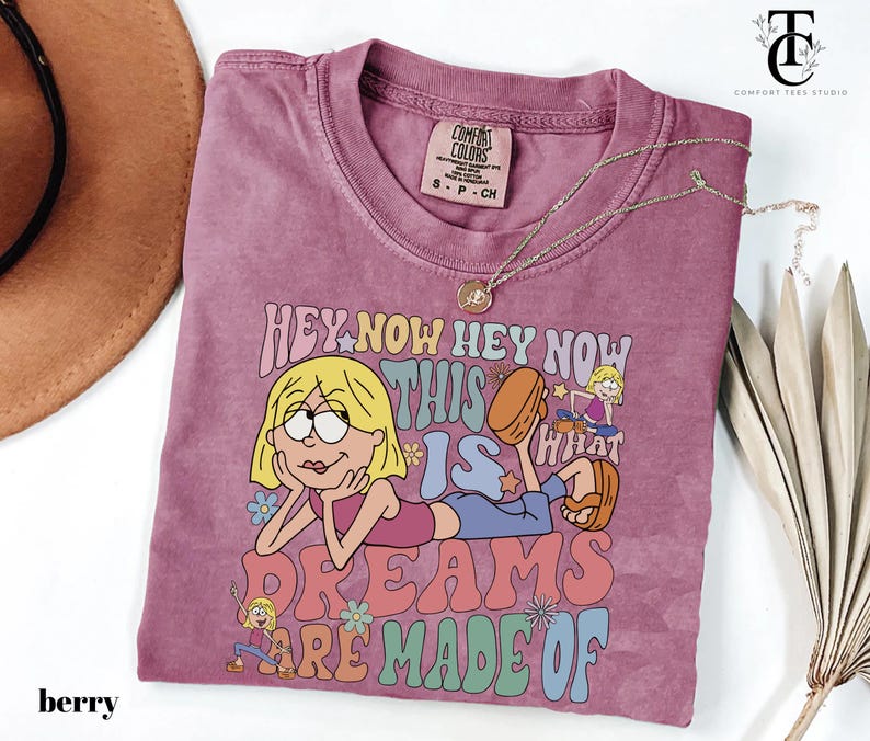 May include: Berry-colored t-shirt with a graphic of a cartoon character and the text "HEY NOW HEY NOW THIS IS WHAT DREAMS ARE MADE OF." The shirt has a crew neck and short sleeves. A gold necklace is draped across the shirt.