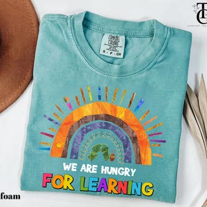 Peut inclure: T-shirt vert d'eau avec un motif arc-en-ciel coloré et le texte "WE ARE HUNGRY FOR LEARNING". Le t-shirt est en matière douce et porte une étiquette Comfort Colors.
