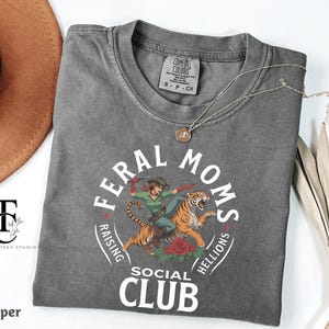 May include: A gray t-shirt with a white graphic of a woman riding a tiger. The graphic includes the text "Feral Moms Social Club" and "Raising Hellions".