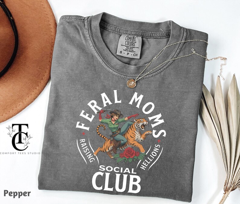 May include: A gray t-shirt with a white graphic of a woman riding a tiger. The graphic includes the text "Feral Moms Social Club" and "Raising Hellions".