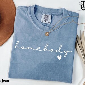 May include: A blue jean t-shirt with the text "homebody" in white script with a small white heart underneath. The shirt has a distressed look and is folded in half.
